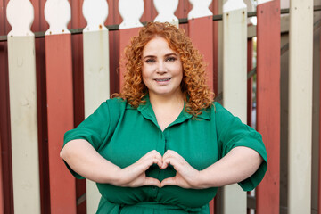 Happy Smiling Plus Size Woman with Curly Red Hair in Green Dress looking at camera, Making Heart Shape gesture with Fingers Outdoors Against Bright Colorful Background