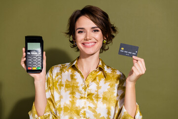 Confident young woman demonstrating credit card and payment terminal in elegant blouse against khaki backdrop