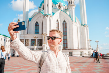 A young male tourist takes a selfie with a smartphone against the background of a Kul Sharif mosque on a cloudy day, Kazan, Russia