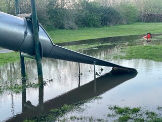Flooded playground with grey metal slide leading to huge puddle and fallen traffic cone.
