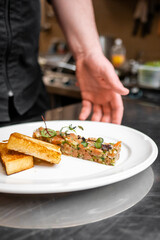A chef’s hand garnishing a plate of tartare with toast in a professional kitchen. The close-up shot highlights the culinary artistry and attention to detail in food presentation