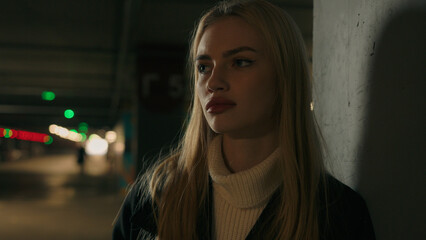 Close up pensive unhappy lost Caucasian young girl female lady businesswoman woman in underground car parking dark shelter evening outside wait taxi looking around upset client consumer alone in night
