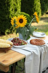 Vase with sunflowers, grapes, pie and corncobs on wooden table in garden