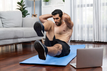 Athletic and sporty man doing crunch on fitness mat while follow online home workout exercise instruction for fit physique and healthy sport lifestyle at home. Online gaiety home exercise video.