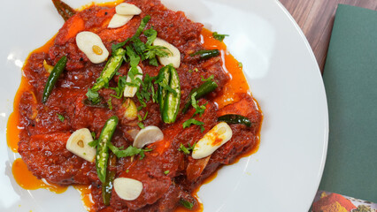 A spicy, rich dish showcased on a white plate. the dish includes tender meat, coated in a deep red tomato-based sauce, garnished with sliced garlic, fresh green chilies, and aromatic herbs.