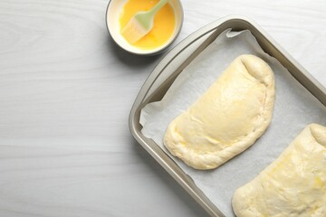 Uncooked calzone pizza in baking dish and egg yolk on white wooden table, top view. Space for text