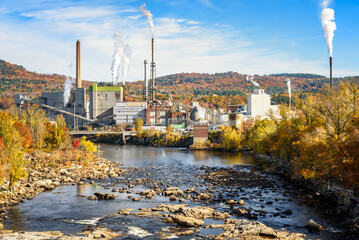 Paper mill with high chimneys belching out colums of white smoke on the bank of a river  in a forested mountain landscape in autumn © alpegor