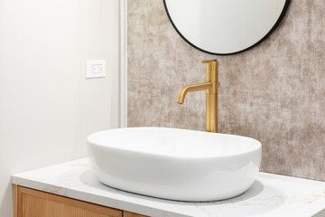 A bathroom sink detail with a gold faucet, white vessel sink on a white marble countertop, wood...