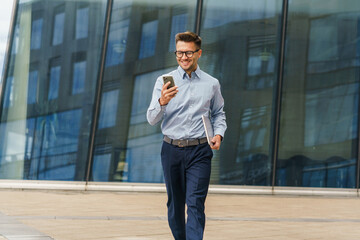 Business professional walking outside while checking smartphone and carrying a notebook in urban setting