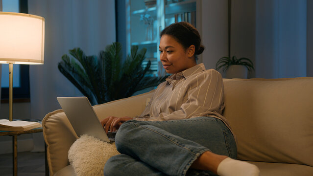 African American woman girl relax on comfortable couch shopping online night time social media addict smiling ethnic female freelancer studying working from home order delivery use laptop application - Powered by Adobe