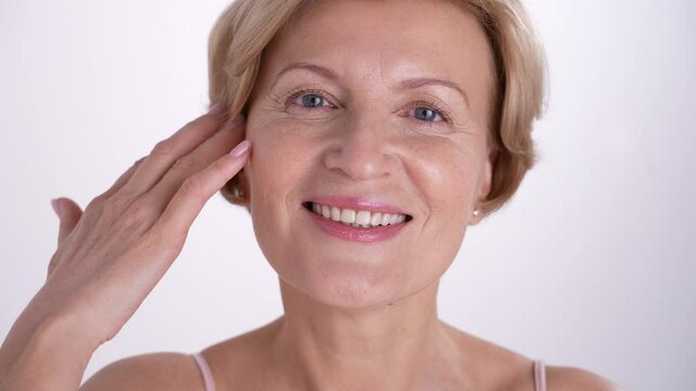 Beautiful woman in her 50s gently touches her radiant face and neck, enjoying her reflection in the mirror. Skincare for aging skin.