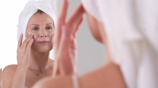 A beautiful mature woman in towel enjoying skincare rituals after shower. Smiling lady applying moisturizing cream. Anti-aging skincare