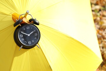 Alarm clock and yellow umbrella on dry leaves in park, closeup. Space for text