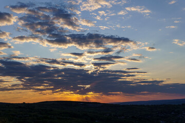 A vibrant sunset casts warm hues across fluffy clouds, creating a breathtaking view over gentle hills in the countryside during evening hours.