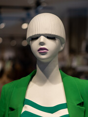 A mannequin showcases current fashion trends in a bright green blazer and a striking bobbed hairstyle. The indoor setting is lively with blurred backgrounds of clothing and accessories.