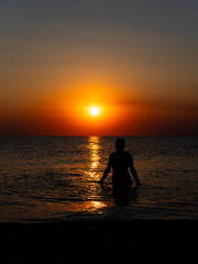 A figure stands in shallow water, gazing at the sunset. The sky displays hues of orange and yellow as the sun dips below the horizon, reflecting on the calm ocean surface.