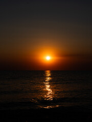 A stunning sunset paints the sky orange and gold as the sun descends beyond the horizon, casting shimmering reflections on the calm ocean water during early evening.