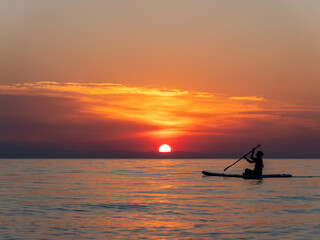 A person paddles on calm waters as the sun sets, creating a stunning silhouette. The vibrant colors of pink, orange, and purple fill the sky, enhancing the serene atmosphere.