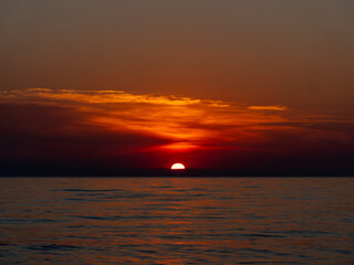 A breathtaking sunset unfolds over the tranquil ocean, with warm orange and red hues illuminating the sky as the sun descends toward the horizon. Soft waves reflect the colors beautifully.