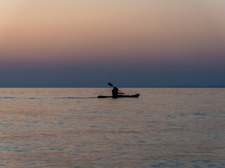 Naklejka premium A person paddles gently across tranquil waters as the sun sets, casting warm orange and soft blue tones across the horizon. The peaceful moment captures the essence of evening solitude.