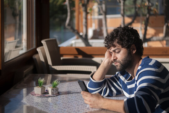 Young man with curly hair using his phone at home. He is looking upset. - Powered by Adobe