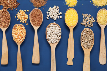 Different types of cereals, seeds and legumes in spoons on blue wooden table, flat lay