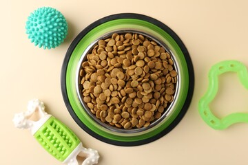 Dry pet food in feeding bowl and toys on beige background, flat lay