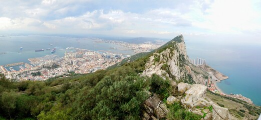 Panoramic view from the top of the Rock of Gibraltar, showcasing the city, marina, Mediterranean Sea, and distant Spanish coastline under a cloudy sky