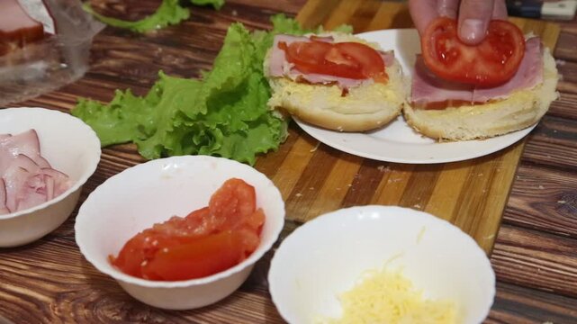 Coocking fastfood at home. Human hands making sandwich from round baked bread, bun, green salad leaves, red tomato slices, meat, bief. Homemade burger. High quality FullHD footage