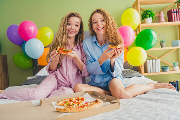 Full size photo of two attractive young women sit bed eating pizza dressed sleepwear outfit wake up cozy day light bedroom home interior