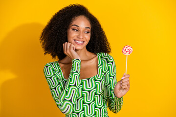 Charming young woman with afro black curly hair in green blouse smiling at lollipop against bright yellow background