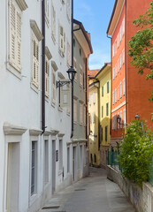 Trieste, Italy - June 27, 2024: Narrow Via dei Capitelli going down at intersection with Via Pozzo di Crosada. Colorful painted facades of residential houses with some green foliage