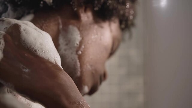 Close-up of young African American woman cleaning herself with soapy sponge from shoulder to neck relaxing in shower. Adult afro black female doing daily hygiene and body care alone in bathroom 