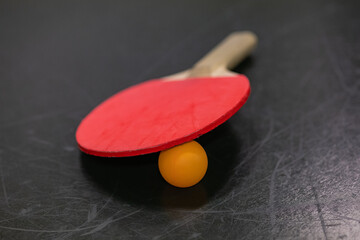 Red table tennis paddle placed over orange ping pong ball on a scratched black table surface....
