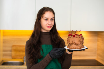 A portrait of a smiling female pastry chef holding a chocolate cake decorated with cherries.