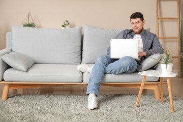 Fototapeta premium Young man sitting on sofa and using laptop in living room