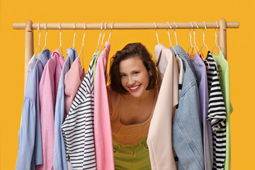 Beautiful young woman near rack with different clothes on yellow background