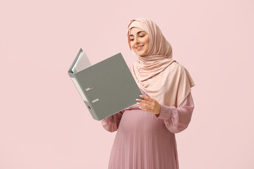 Beautiful pregnant Arabian businesswoman in hijab with folder on pink background