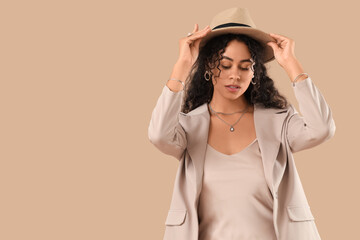 Beautiful young African-American woman with stylish jewellery and wicker hat on beige background