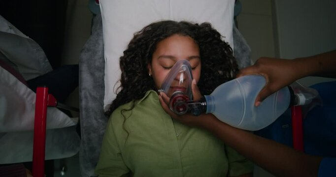 The patient is lying on a gurney with her eyes closed. The nurse gives her air through an oxygen mask. View from above. Emergency resuscitation