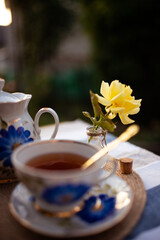 Closeup of a yellow rose in a tiny glass vase. A table is served and decorated with a beautiful porcelain set of dishes decorated with blue flowers. Breakfast in the garden on a sunny day. 