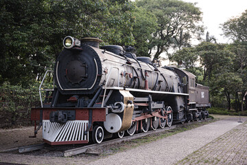 An antique steam locomotive displayed outdoors, showcasing intricate details and historic design. The train's metal structure contrasts with a softly blurred background, emphasizing its vintage charm.