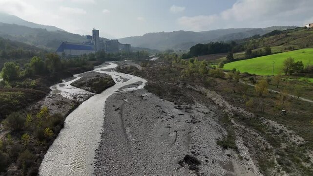 River flowing through valley with rocks and vegetation in autumn and the unicem cement factory in the background, Arda River in Val Tolla, Lugagnano D'Arda, Italy, slow motion drone flying forward