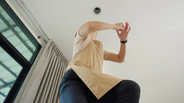 Low-angle view of Asian woman wearing an apron standing on a ladder and changing a light bulb of a downlight in a ceiling fixture indoors.