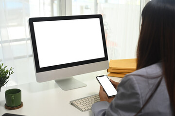 Side view of female employee holding smartphone with empty screen sitting in front of computer desktop