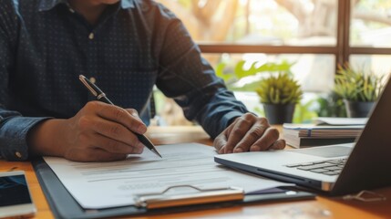Fototapeta premium Individual Writing on Documents at a Wooden Desk with Laptop and Green Plants in a Brightly Lit Office Environment for Business or Academic Purposes