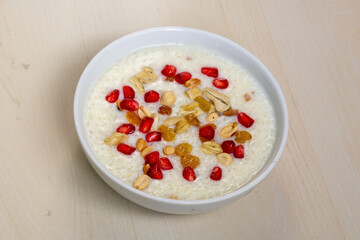Homemade tasty payesh or payasam, served in a white bowl on a wooden background. Cooked with milk, sugar or jaggery, and rice, it's garnished with pomegranate, cashew nuts, raisins, and peanuts.