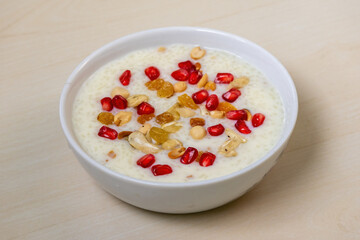Delicious homemade payesh or payasam, served in a white bowl on a wooden background. Cooked with milk, sugar or jaggery, and rice, it's garnished with pomegranate, cashew nuts, raisins, and peanuts.