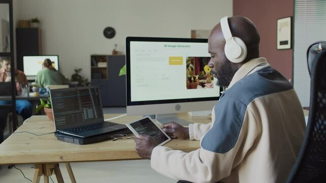 Medium side shot of black male IT engineer writing AI software using laptop and digital tablet, then running it on desktop computer to check performance, then looking at code for bugs and errors