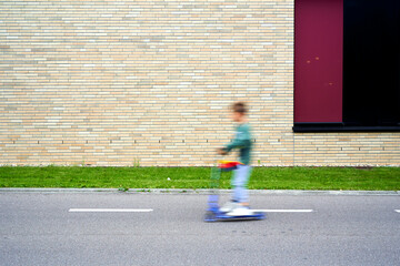 A child on a scooter speeds past a brick wall, blurring motion against static urban backdrop.